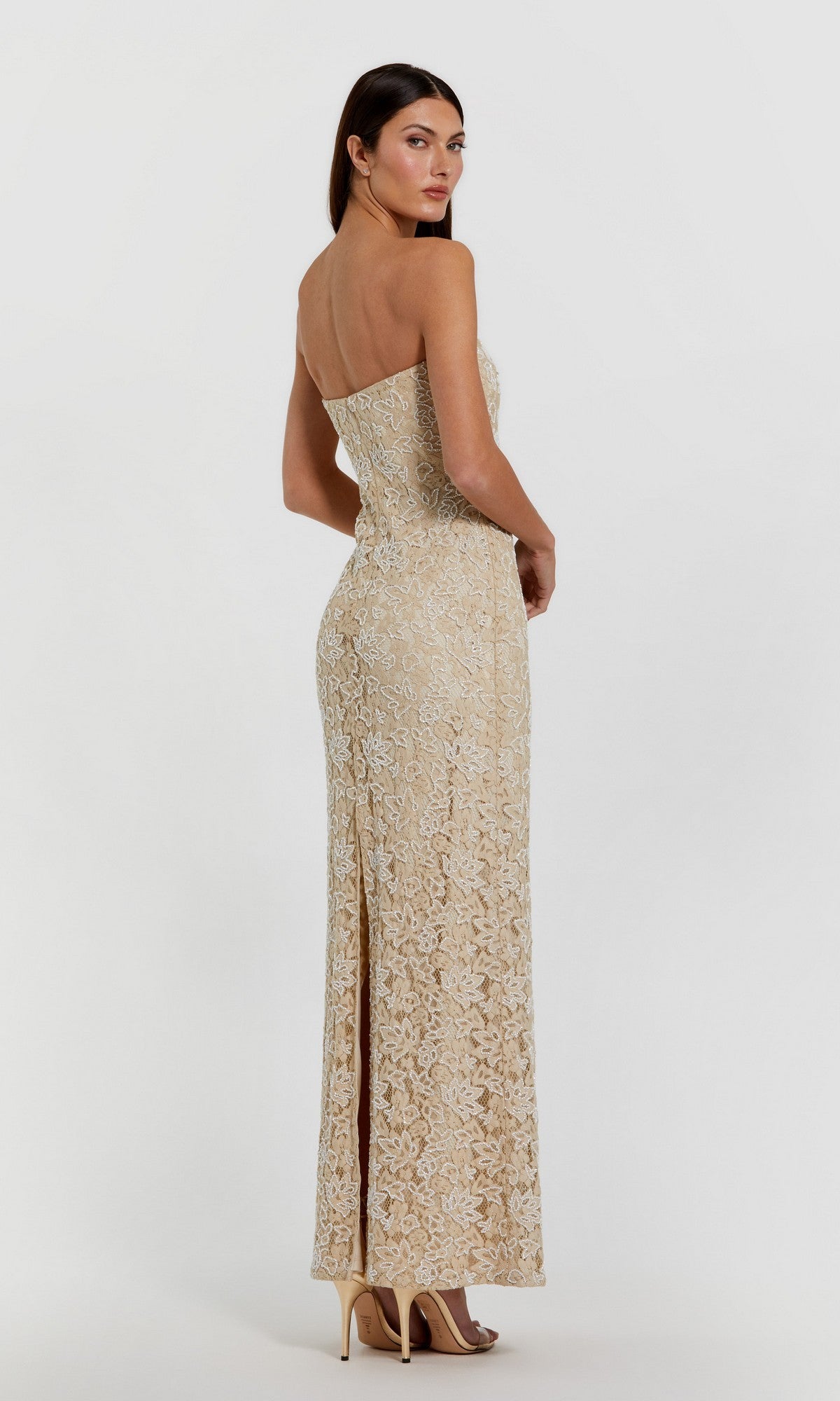 A woman in the Long Formal Dress: Mac Duggal 24067, a strapless beaded lace column gown with a back slit, and high heels stands against a plain white background.